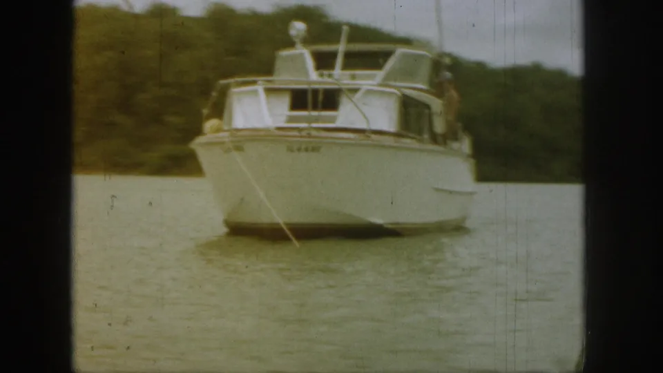 1961 Michigan 8mm frame of a white motorboat angled across calm lake water, with trees behind and a tow line visible.