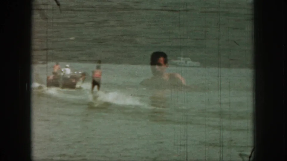 1961 Michigan lake footage showing a motorboat and waterskier with a superimposed swimmer or floating man at right.