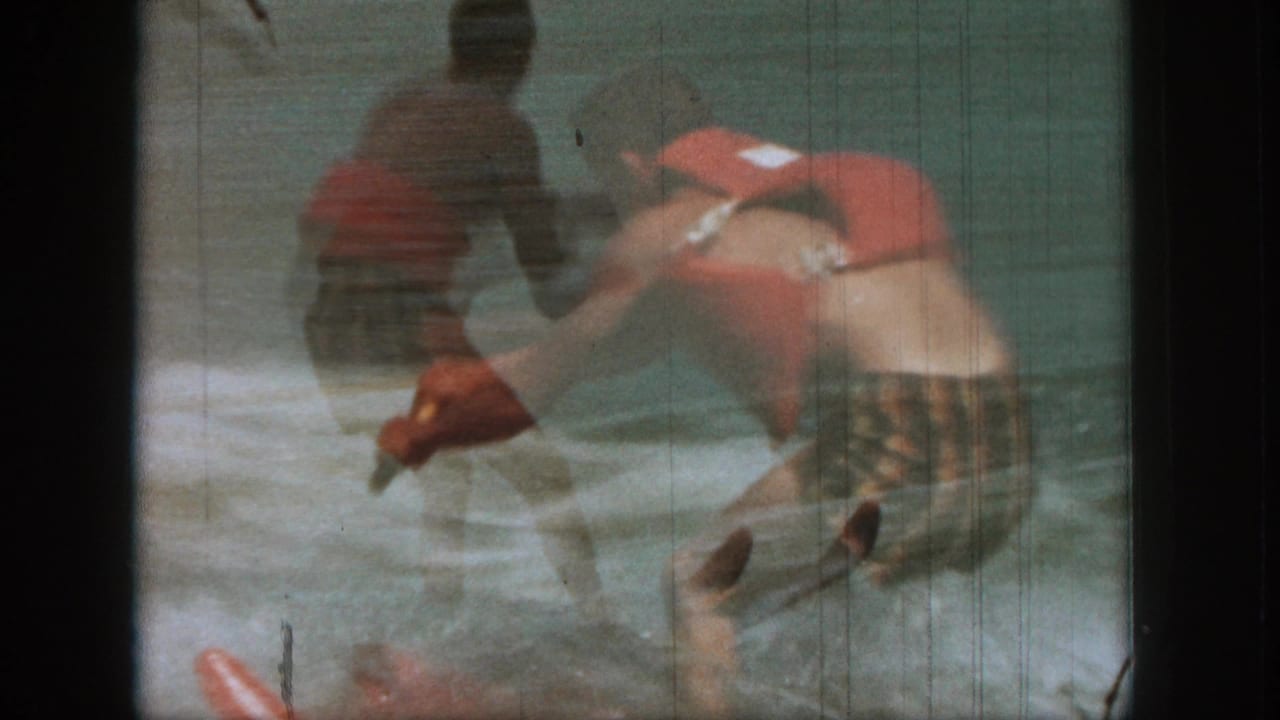 1961 Michigan 8mm frame of a man in a red life vest and striped trunks leaning forward while water skiing on a lake.