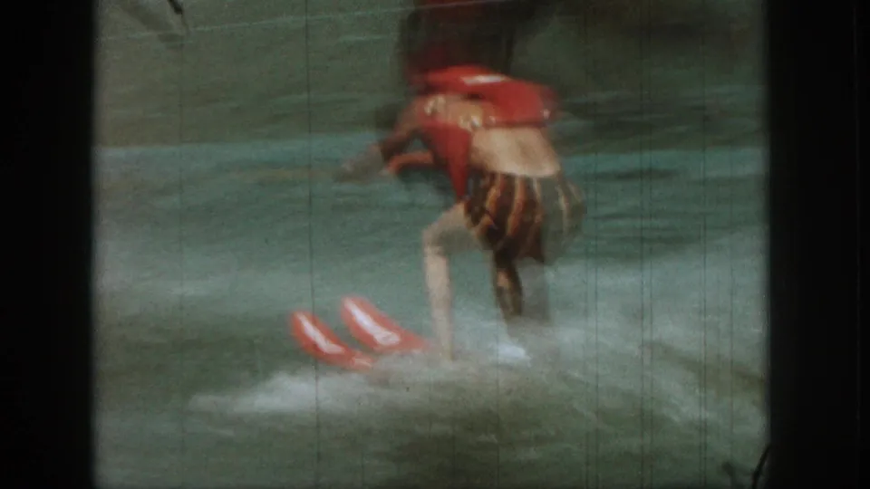 The skier moves across the wake in red safety gear and striped trunks, with water spray and blurred motion on the lake.