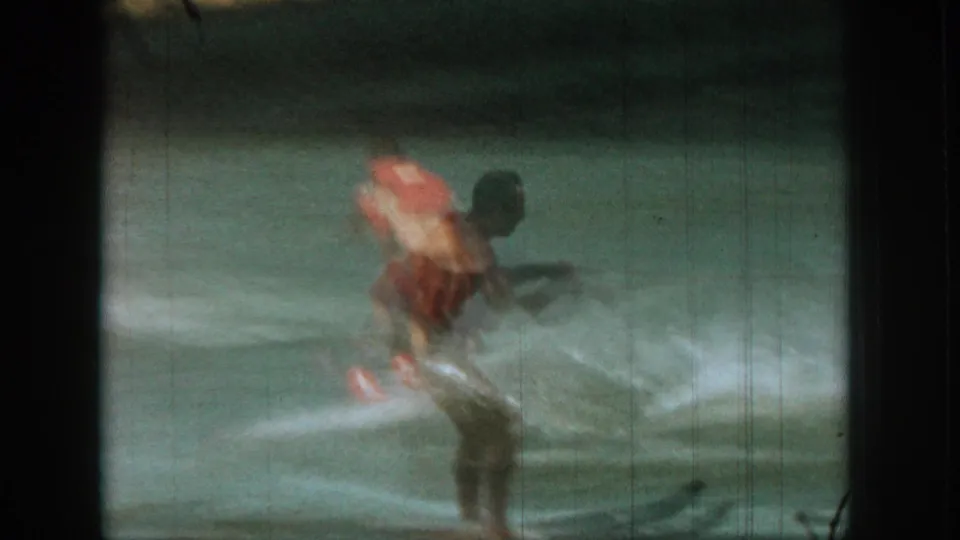 A home-movie view from the boat shows the skier balancing on red skis while spray rises around his legs.
