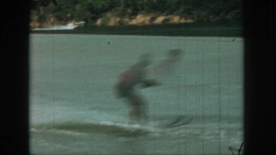 A motion-blurred skier crosses the lake, with a wooded shoreline and small distant boat visible in the background.