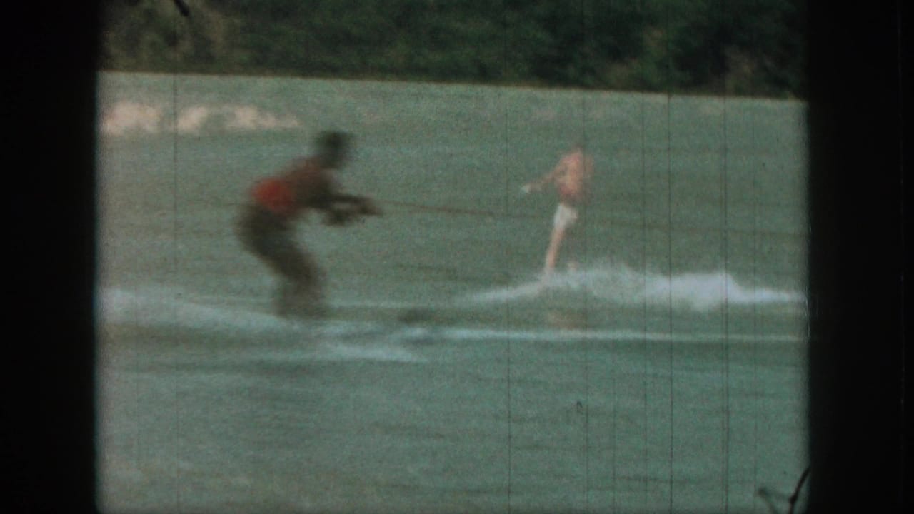 Water skier in a red top crosses a river while a motorboat follows in the background.