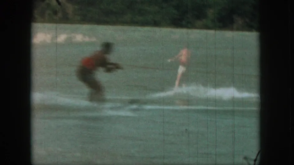 1961 Michigan home-movie frame showing a red-shirted water skier on calm water with a towing motorboat behind him.