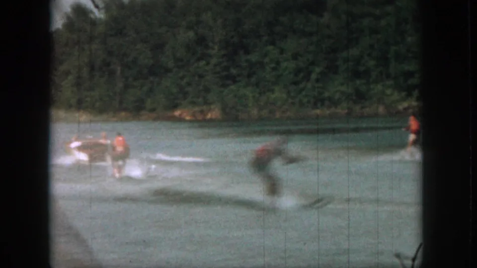 1961 Michigan home-movie frame with a motorboat towing a skier across a calm river near trees.