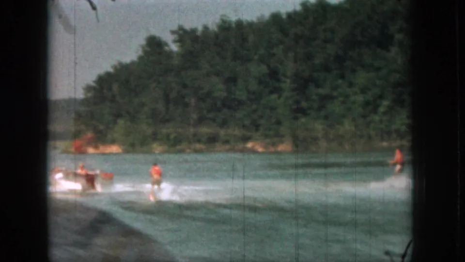 Wide 1961 Michigan home-movie frame of water skiing on a river, with the boat and distant figures against a wooded shoreline.