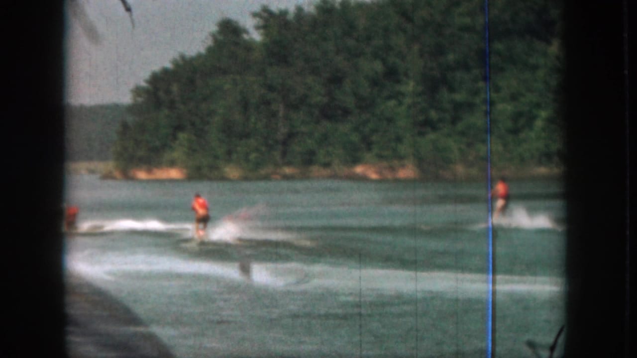 1961 Michigan 8mm frame showing a water skier in an orange life vest moving across a lake, with dark film edges and trees beyond.