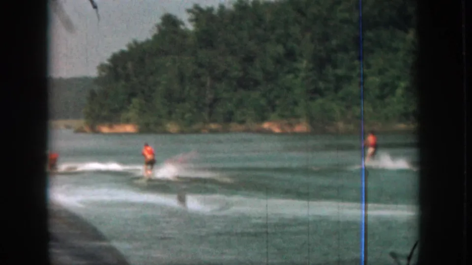 1961 Michigan 8mm frame showing a water skier in an orange life vest moving across a lake, with dark film edges and trees beyond.