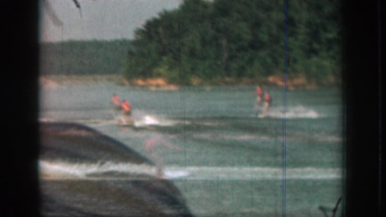 1961 Michigan lake scene with two orange-vested water skiers following a tow boat, framed by dark 8mm borders and visible scratches.