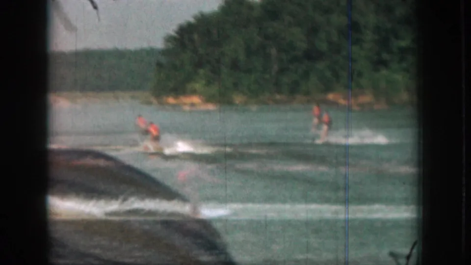 1961 Michigan lake scene with two orange-vested water skiers following a tow boat, framed by dark 8mm borders and visible scratches.