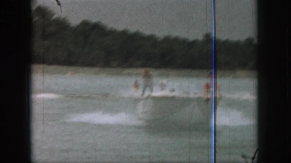 1961 Michigan 8mm frame showing two distant adult water skiers pulled behind a small boat on a tree-lined lake.