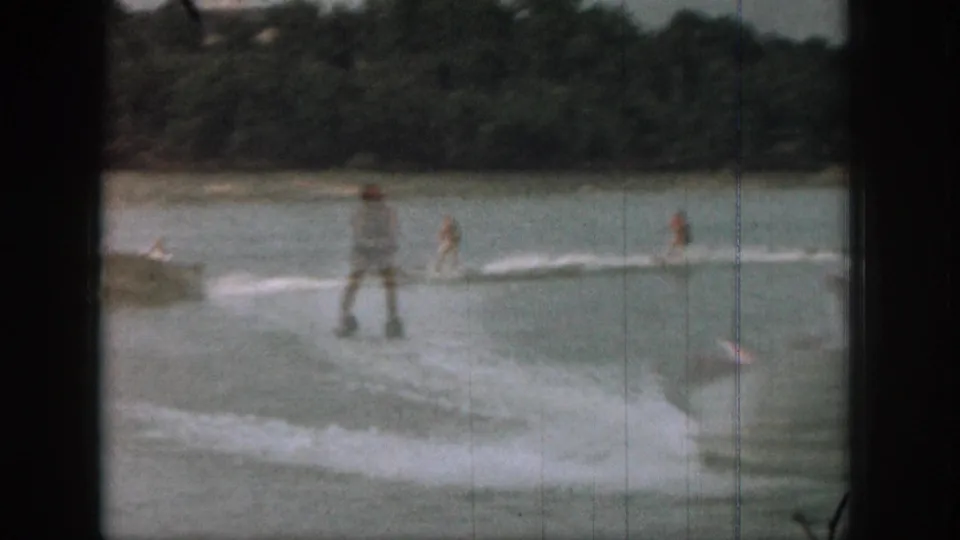 Adult water skiers and a small boat cross a Michigan lake in a soft, grainy 1961 8mm home-movie frame.