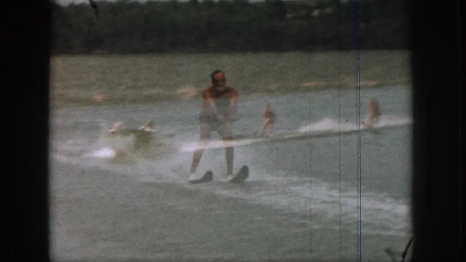 Closer 1961 Michigan lake view of an adult male water skier in swim trunks, with boat wake and another skier behind.