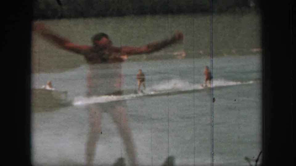 1961 Michigan 8mm frame with a closer adult water skier raising his arms as a boat and another skier pass behind him.