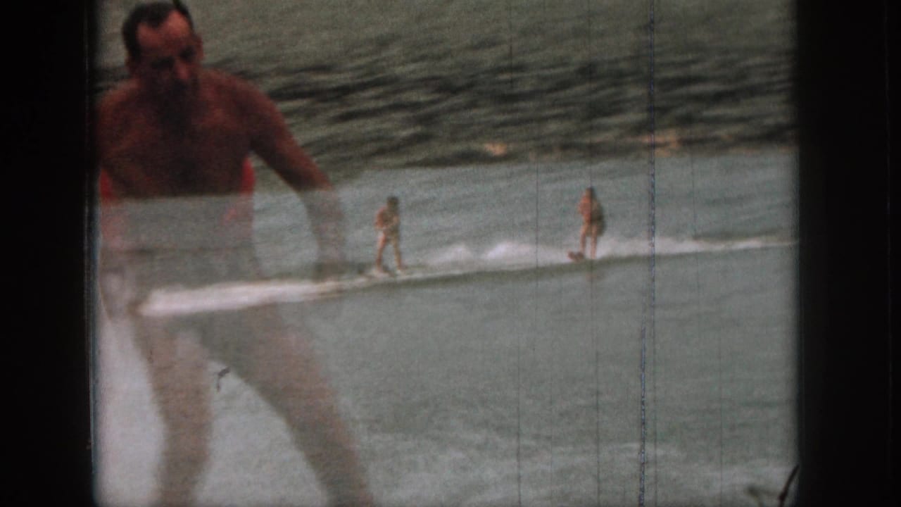 1961 Michigan home-movie frame with a water skier in the distance and an adult in swim trunks partly foregrounded near the lake.