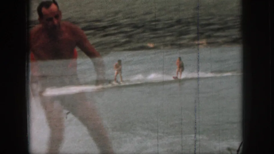 1961 Michigan home-movie frame with a water skier in the distance and an adult in swim trunks partly foregrounded near the lake.