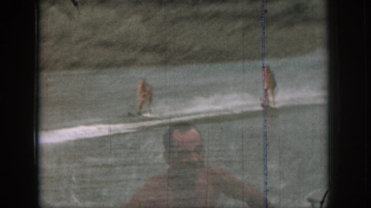 Two water skiers ride across a lake with a swimmer or wader low in the foreground.