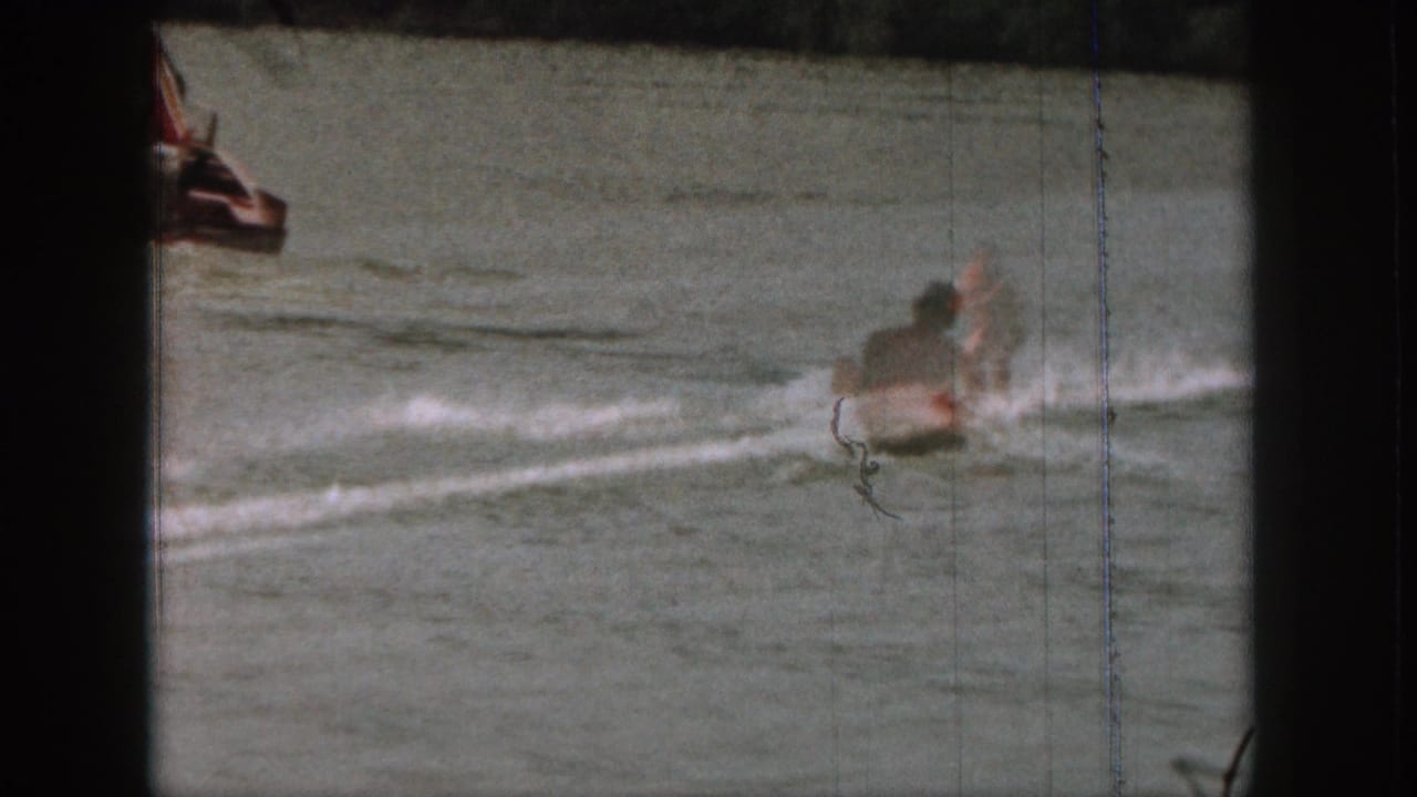 1961 Michigan home-movie frame with a water skier throwing spray beside a lake surface and a partial boat edge at left.