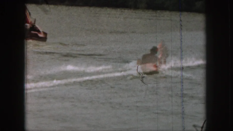 1961 Michigan home-movie frame with a water skier throwing spray beside a lake surface and a partial boat edge at left.