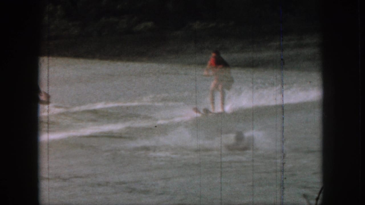 1961 Michigan 8mm frame showing a water skier in red swimwear gliding across the lake with shoreline vegetation beyond.