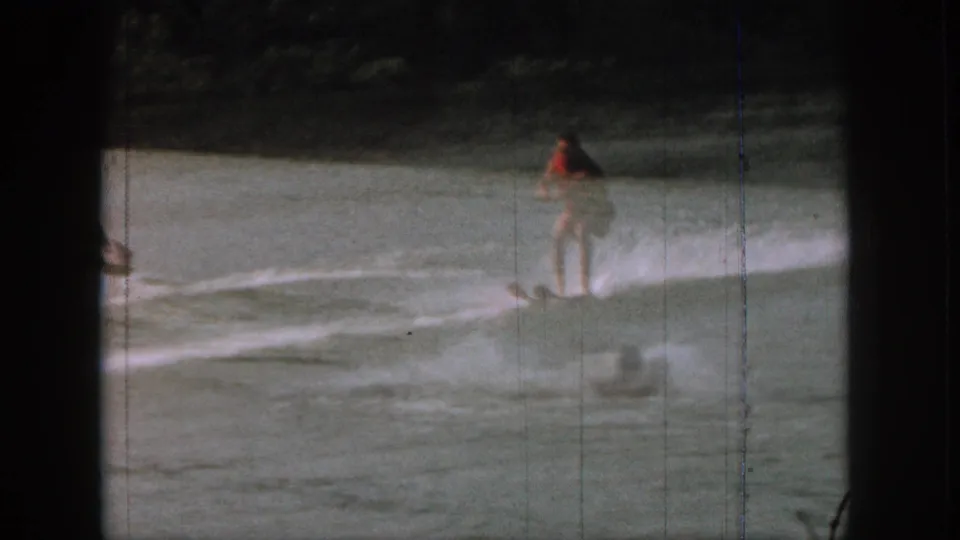 1961 Michigan 8mm frame showing a water skier in red swimwear gliding across the lake with shoreline vegetation beyond.