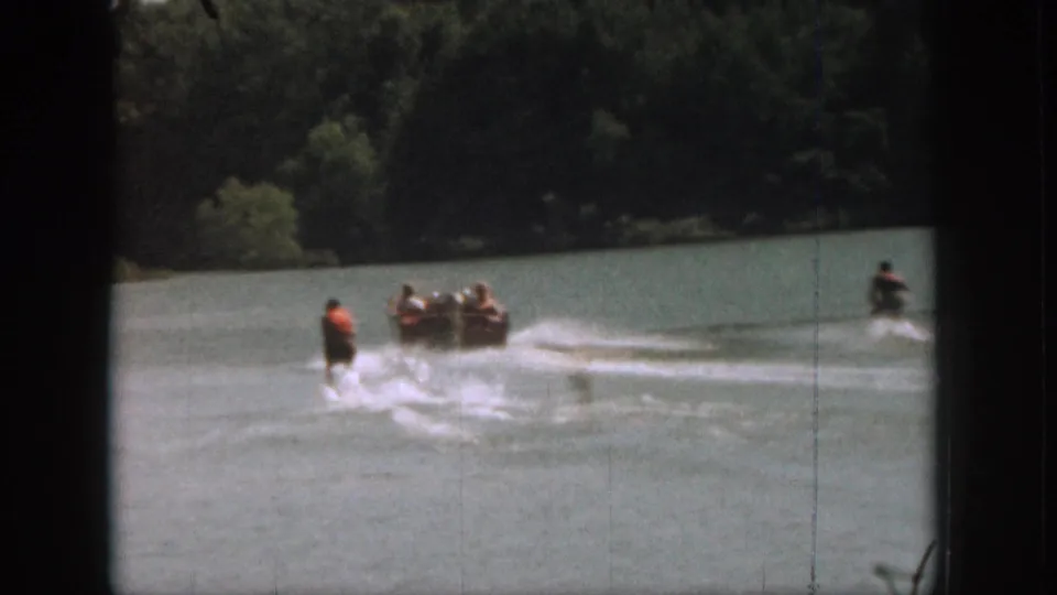 1961 Michigan home-movie frame showing a motorboat pulling skiers across a tree-lined lake, with one skier farther to the right.