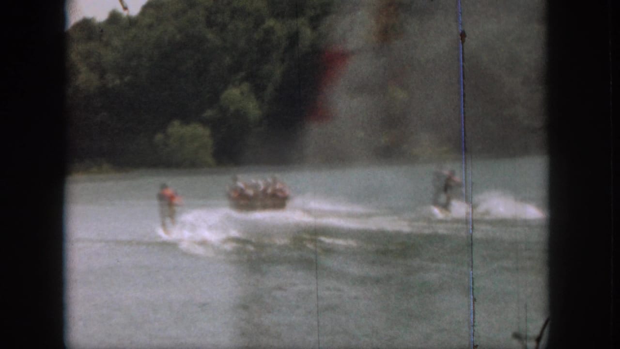 Water skier crosses a lake behind a motorboat with wooded shoreline in the background.