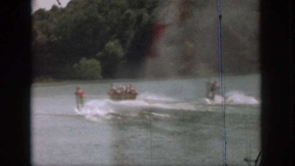 1961 Michigan home-movie frame showing a man water skiing behind a small motorboat on a wooded lake.