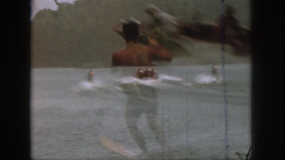 Home-movie frame from 1961 Michigan with a foreground adult figure and a water skier moving behind a boat on the lake.