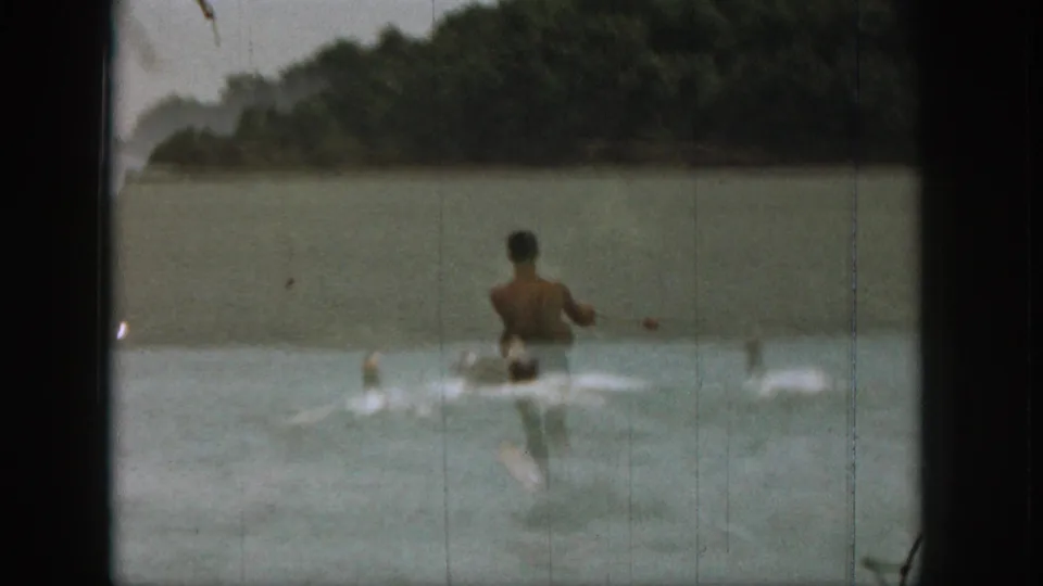 1961 Michigan 8mm frame showing a rear view of a man water skiing across a quiet lake bordered by trees.