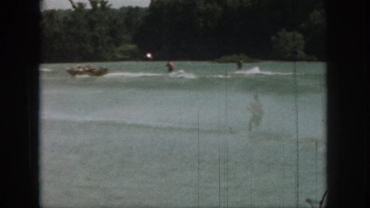 Wide 1961 Michigan home-movie view of a skier following a small motorboat across a lake bordered by trees.