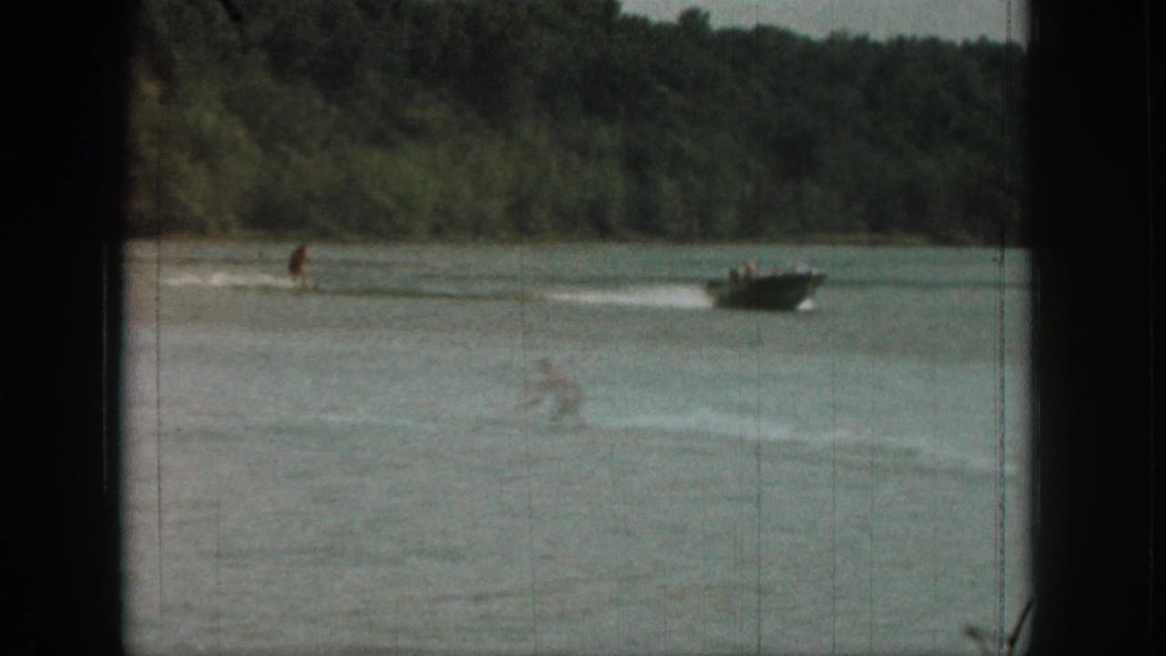 Distant 1961 Michigan lake frame with a water skier, towing boat, white wake, and dense tree-lined shoreline.