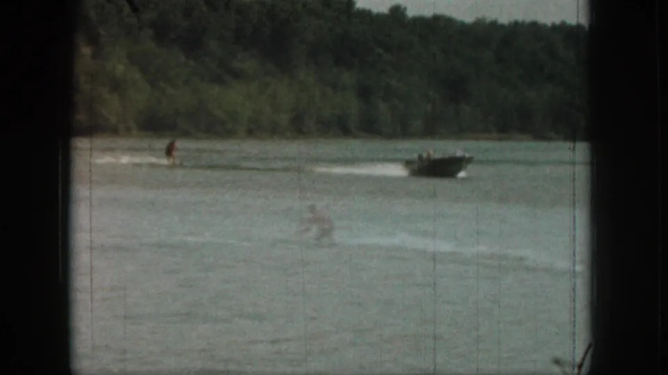 Distant 1961 Michigan lake frame with a water skier, towing boat, white wake, and dense tree-lined shoreline.