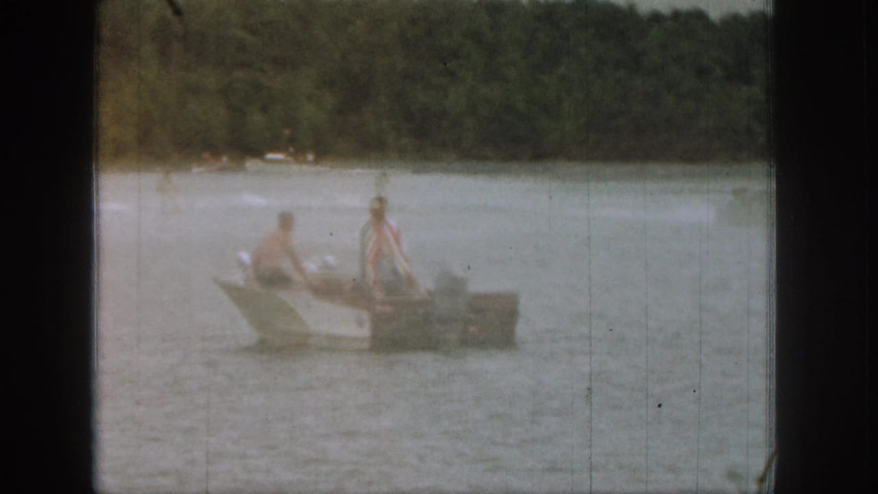 Small boat on a lake with two people aboard near faint wake spray.