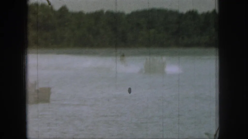 1961 Michigan 8mm frame showing a distant boat wake and spray across a wooded lake, with film gate edges visible.