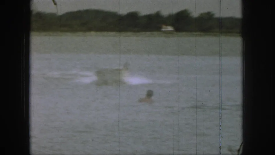 1961 Michigan home-movie frame showing a distant swimmer in open lake water with a white water-ski wake and wooded shoreline behind.
