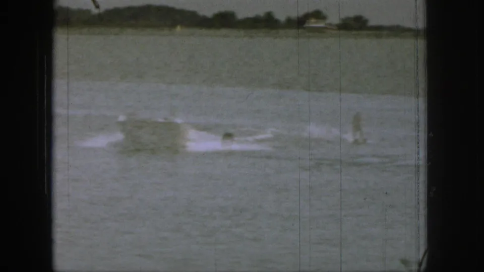 1961 Michigan home-movie frame with a water skier or swimmer activity in the distance, white wake on the lake, and a tree-lined shore.