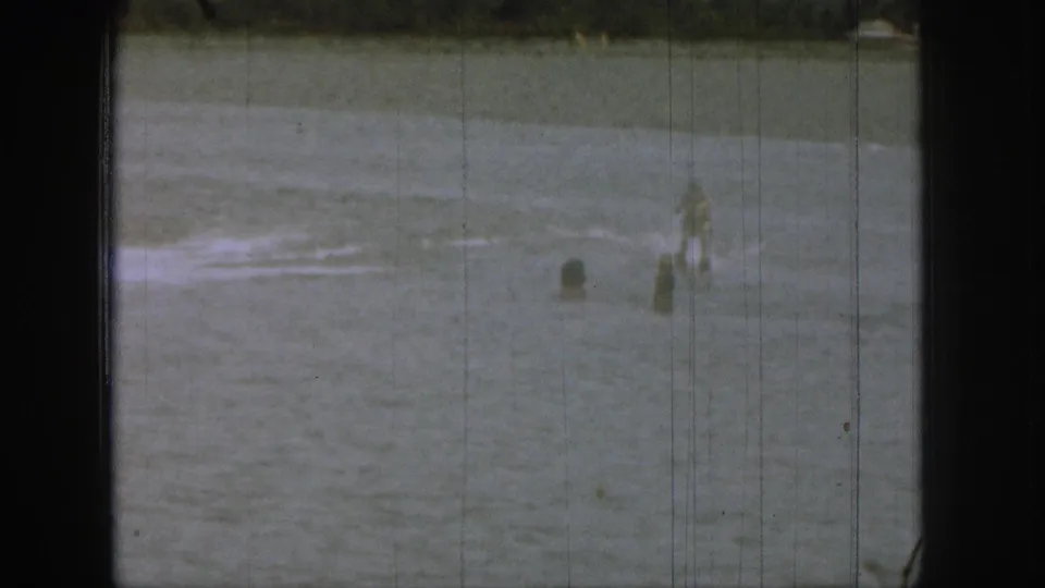 1961 Michigan 8mm frame showing swimmers spread across a lake with wake on the water and a low shoreline in the background.