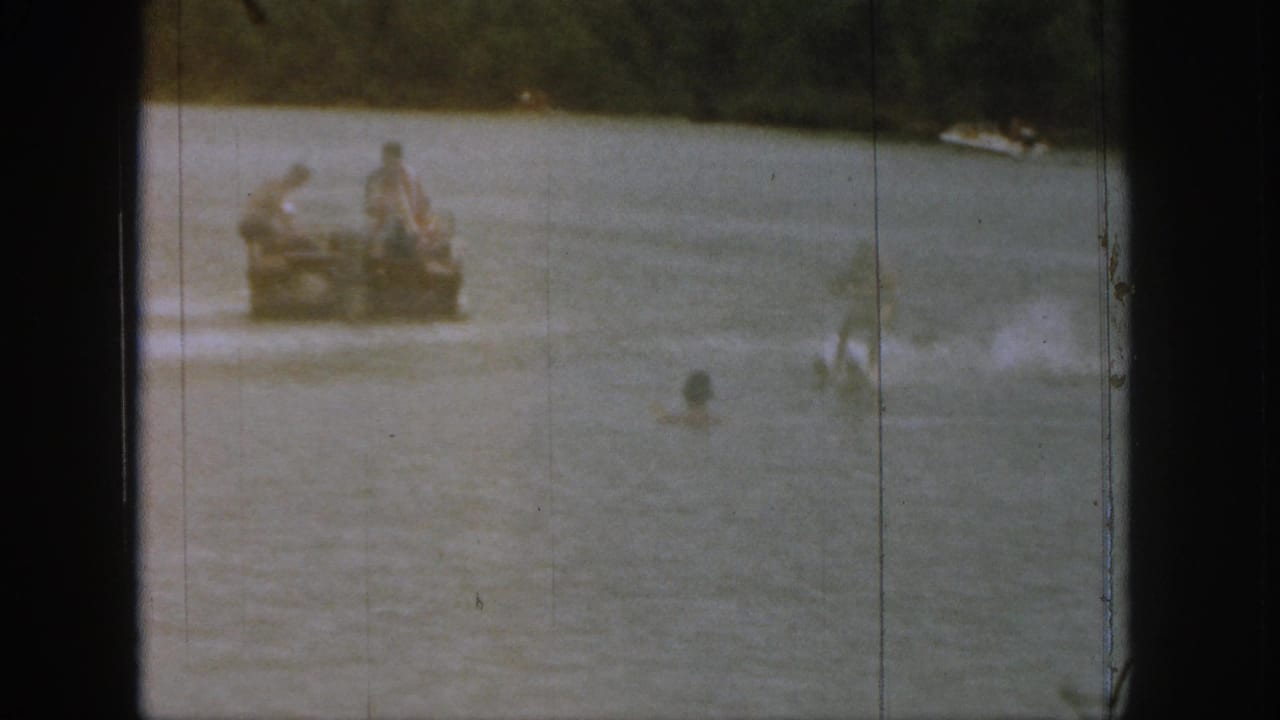 1961 Michigan home-movie frame showing people seated in a small motorboat on a lake with swimmers nearby and wooded shore beyond.
