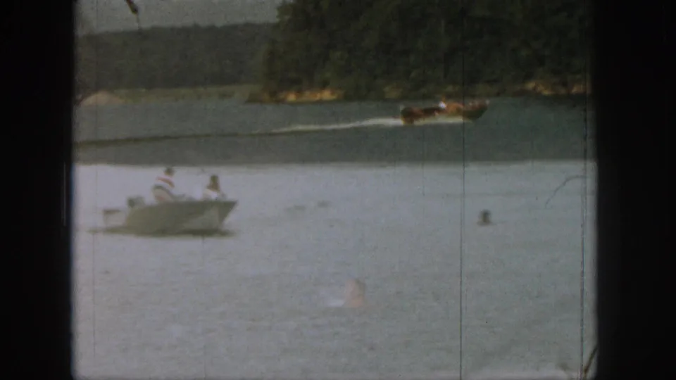 1961 Michigan 8mm frame showing a small rowboat with people aboard, swimmers nearby, and a motorboat farther across the lake.
