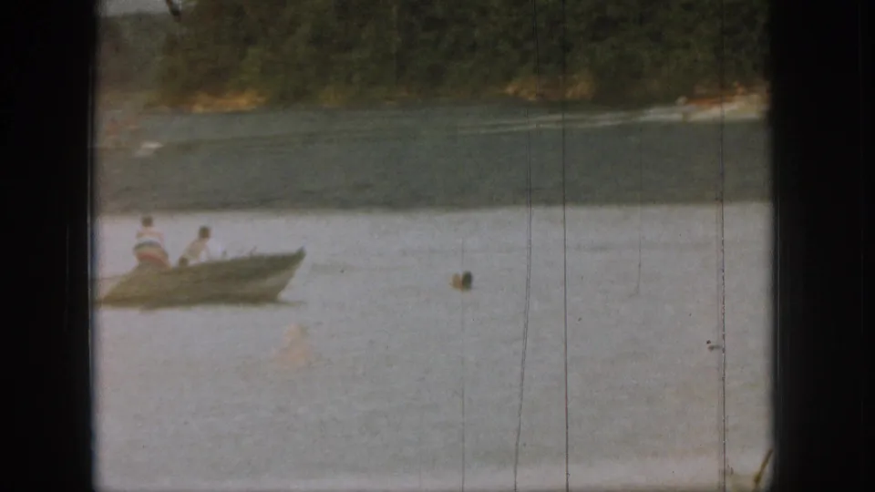 1961 Michigan home-movie frame with a small rowboat, people seated inside, a nearby swimmer, and a wooded lakeshore in the distance.