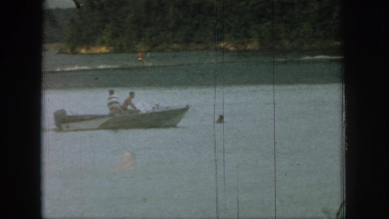 Small motorboat with two passengers on a lake, with a distant water skier and wooded shoreline.