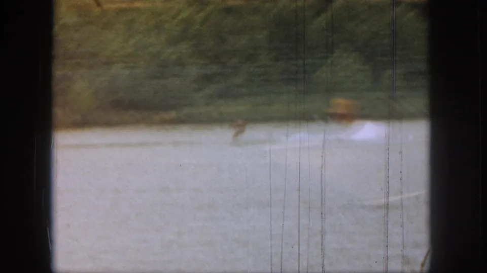 Soft 1961 Michigan lake frame with a blurred foreground figure, pale water surface, and wooded shore in the background.