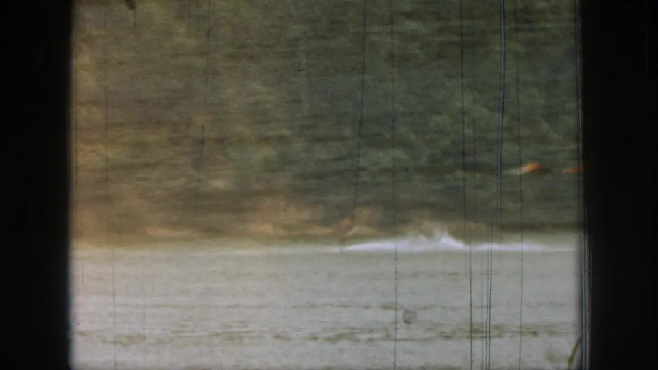 Soft 8mm view through a boat window, with lake spray and a tree-lined Michigan shore visible as the skier moves out of frame.