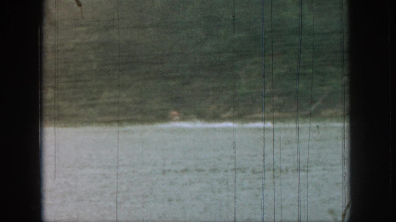 1961 Michigan 8mm frame showing a small distant figure low in lake water with a dark shoreline behind.