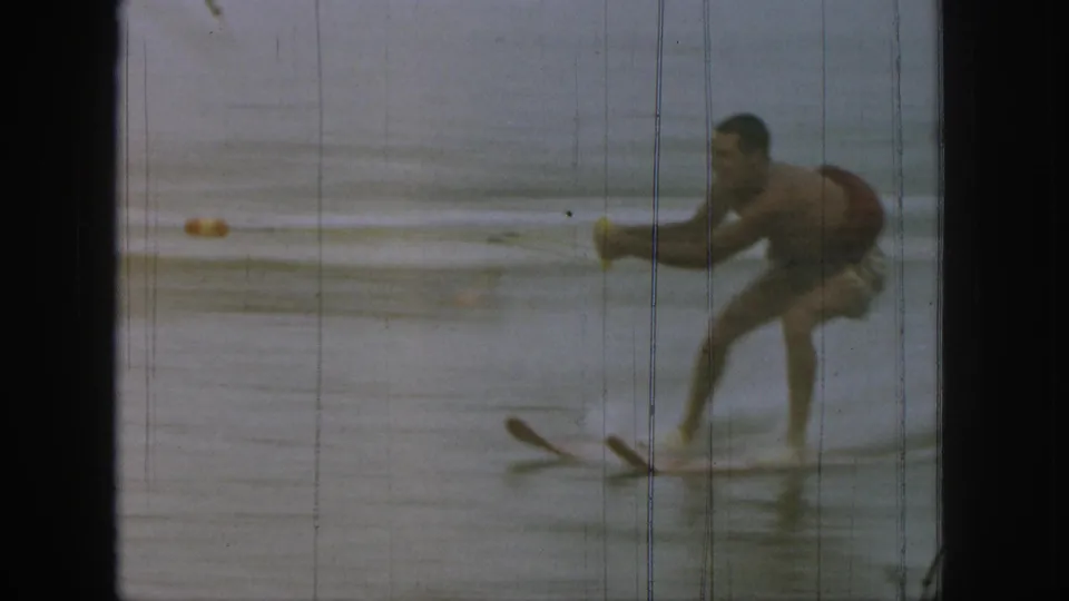 1961 Michigan 8mm frame showing a young water skier bent forward, balancing on paired skis over calm lake water.