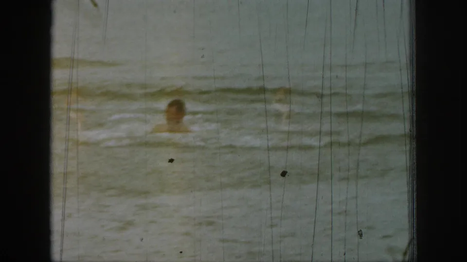 1961 Michigan home-movie frame showing a swimmer's head above rippling water, with faded color and vertical 8mm scratches.