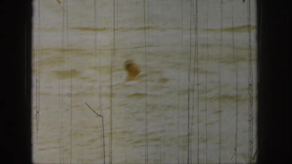 1961 Michigan home-movie frame showing a swimmer in rippling water, with warm fading, heavy scratches, and visible frame edges.