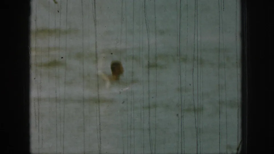 1961 Michigan home-movie frame showing a distant person partly obscured by lake spray and heavy vertical film scratches.