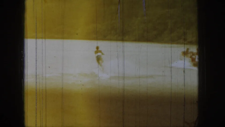 Distant waterboarder crossing a Michigan lake in 1961 home-movie footage, partly obscured by grain, scratches, and soft focus.